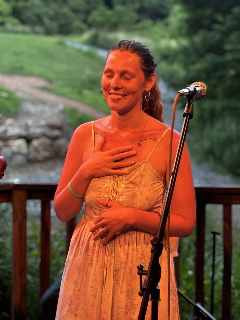 Katt at a microphone at dusk, eyes closed and one hand on her heart