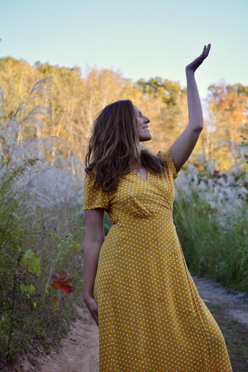 Katt in a yellow dress in an autumn meadow, one hand reaching up to the sky